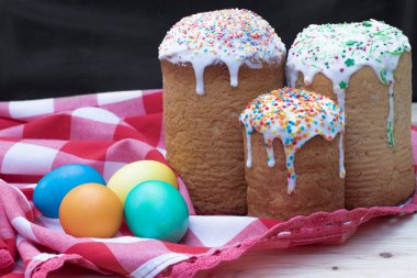 Üç fırınlanmış Paskalya pastası üzerine şeker serpiştirilmiş, dört renkli tavuk yumurtası kırmızı ve beyaz bir bez ve siyah bir arka plan, yakın plan.
