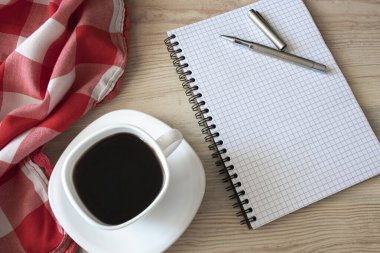 Beyaz fincan koyu siyah kahve, kırmızı-beyaz bez, metal kalem ve ahşap yüzey üzerine not defteri.
