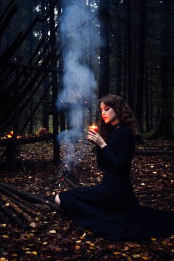 Büyülü cadılar bayramı cadısı, ateşin yanında, ormanda büyüler. Hokkabazlık yapan genç ve güzel bir kadın. Ürkütücü kara büyü ormanı geçmişi. Büyücü. Cadılar Bayramı parti tasarımı