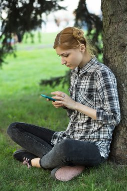 Ağaç parkta oturan ve smartphone kullanarak genç kadın