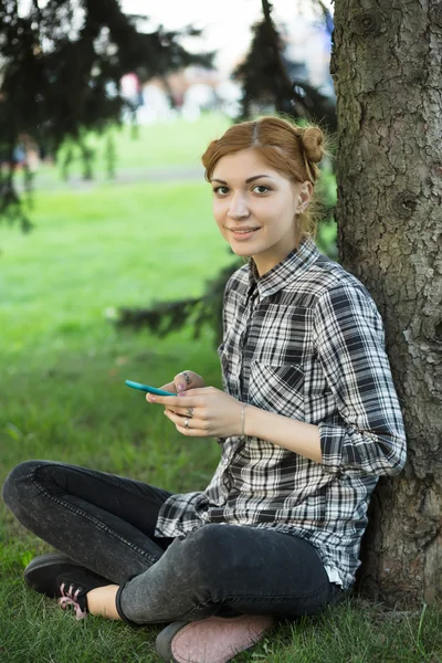Young woman sitting at tree in park and using smartphone — Stock Photo, Image
