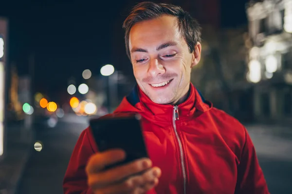 Smiling man in red using mobile phone at the night city street