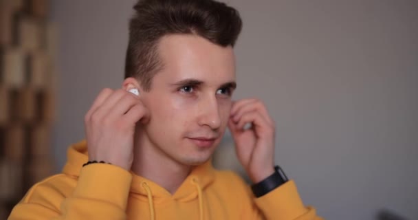 Man in yellow hoodie puts on wireless earbuds into his ears at home — Stock Video