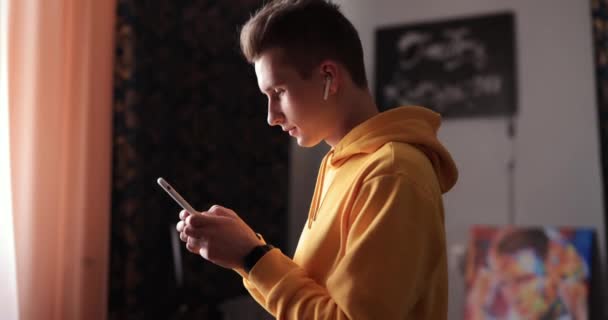 Man in yellow hoodie using smartphone and earbuds at home — Stok video