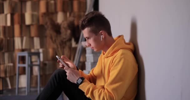 Man in yellow hoodie using smartphone and earbuds at home — Αρχείο Βίντεο