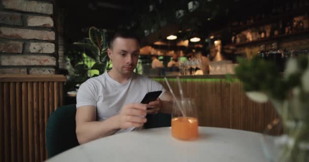 El hombre bebe jugo de frutas en el restaurante mirando en el teléfono inteligente — Vídeo de stock