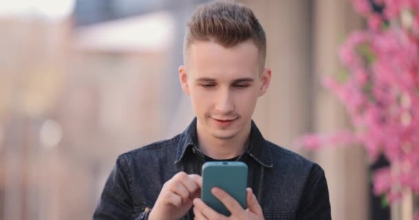 Молодой улыбающийся мужчина, использующий мобильный телефон в весеннем городе — стоковое видео