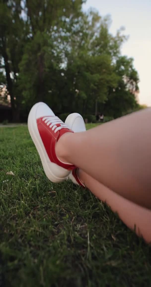 Las piernas femeninas en zapatos de goma rojos descansando sobre una hierba, vídeo vertical — Vídeo de stock