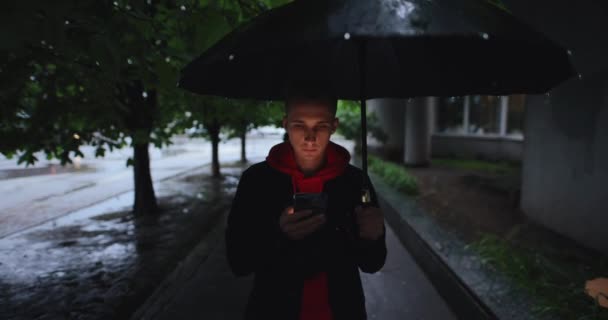 Человек идет по аллее под зонтиком с помощью смартфона — стоковое видео