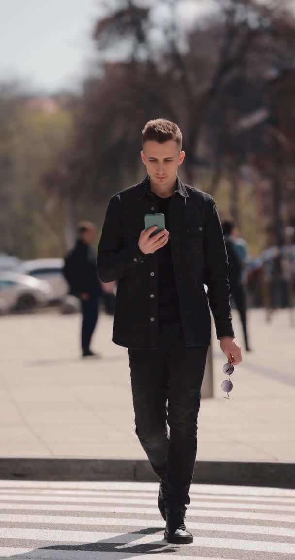 Młody człowiek korzystający z telefonu komórkowego przechodzącego przez ulicę na zebrze, pionowe wideo — Wideo stockowe