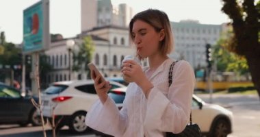 Bir yaz şehrinde yürürken cep telefonuyla mesaj atan bir kadın kokteyl içiyor.