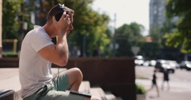 Gülümseyen adam yaz şehrinde otururken büyük kulaklık takar.