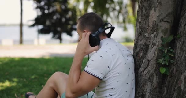 Homem coloca grandes fones de ouvido apoiados na árvore — Vídeo de Stock