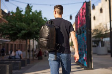 Arkadan bakan adam siyah deri sırt çantasıyla şehirde yürüyor.