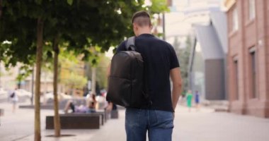 Arkadan bakan adam siyah deri sırt çantasıyla şehirde yürüyor.