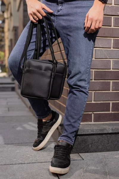 Unrecognizable man holding two messenger bags in his hands leaning at brick wall