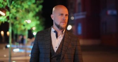 Klasik takım elbiseli kel bir adam gece şehirde yürüyor.