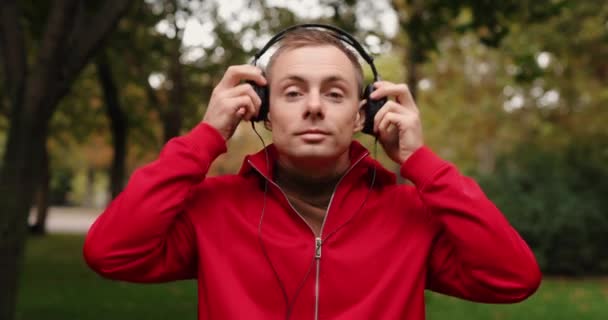 Hombre de chaqueta roja con auriculares en el parque de otoño — Vídeos de Stock