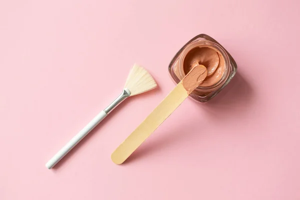 Facial pink clay mask for sensitive skin, wooden stick and make-up brush on pink background with copy space, top view. Skin care beauty delicate detox product minimalistic flat lay