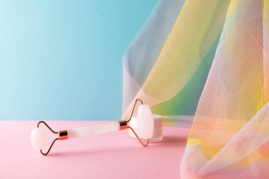 Renkli gökkuşağı organza örtüsüyle mavi arka planda, cilt bakımı için kuvars yüz silindiri ve nem kremi.