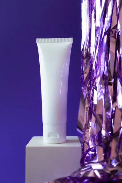 White plastic tube mockup with moisturizer cream, shampoo or facial cleanser and holiday tinsel on cube podium on violet background, vertical — стоковое фото