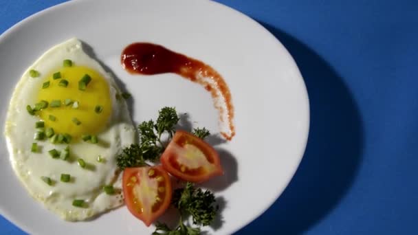 Danie z jajecznicą. Jajka smażone na białym talerzu z pomidorami, ziołami, ketchupem. Niebieskie tło z miejscem na tekst. Śniadanie kawalerskie. — Wideo stockowe