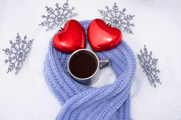 Red hearts and coffee in an old metal mug in the snow wrapped in a hand-knitted lilac scarf. New year\'s composition for Christmas 2021 with Christmas toys in the form of snowflakes.