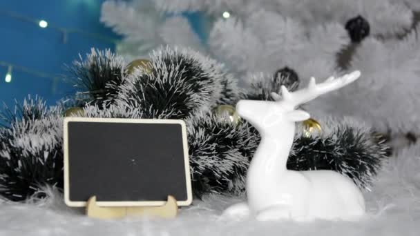 Julbakgrund. Julgran med nyårsträdgård. En vit hjort ligger bredvid en textskylt. Gran grön snötäckt gren ligger i snön. — Stockvideo