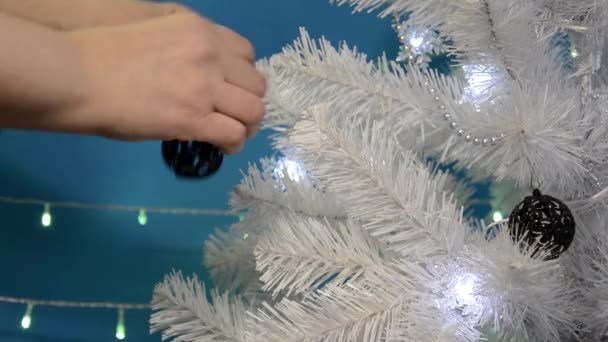 Kerst achtergrond. Close-up handen versieren een witte kerstboom met zwarte visnet ballen. Kerstboom met knipperende lichtjes en kerstspeelgoed op een blauwe achtergrond. Gelukkig Nieuwjaar. — Stockvideo