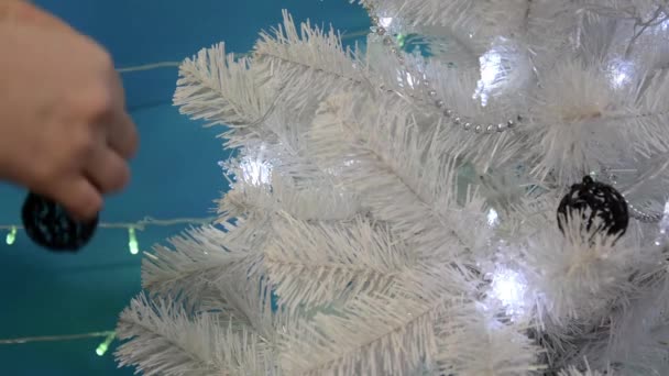 Las manos de cerca decoran un árbol de Navidad blanco con bolas de red negra. Árbol de Navidad con luces parpadeantes y juguetes de Navidad sobre un fondo azul. Fondo de Navidad. Feliz Año Nuevo. — Vídeos de Stock