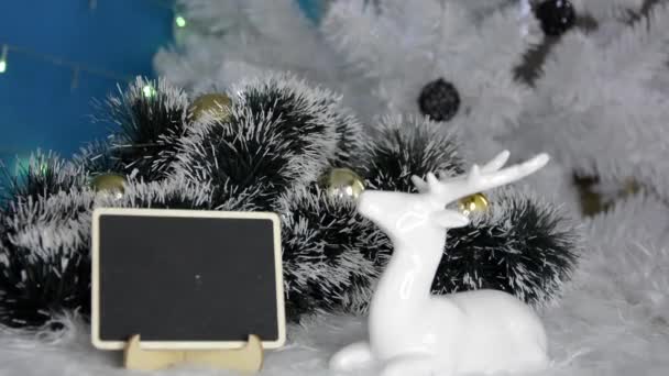 Frohes neues Jahr. Weihnachtlicher Hintergrund. Weihnachtsbaum mit Neujahrsgirlanden. Ein weißes Reh liegt neben einem Schild mit einem Glückwunschtext. Fichtengrüner schneebedeckter Ast liegt im Schnee. — Stockvideo