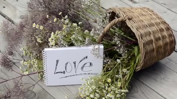 La parola amore su sfondo bianco. Dichiarazione d'amore sullo sfondo di fiori di campo in un cesto di vimini. Prato di erbe fiorite in una giornata di sole. Bouquet panoramico estivo. — Video Stock