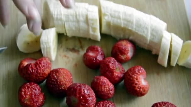 Una mano corta un plátano y una fresa en pedazos. Fondo borroso para la pantalla de salpicadura. La preparación de la ensalada de frutas para la alimentación dietética. Fruta en la cocina. — Vídeos de Stock