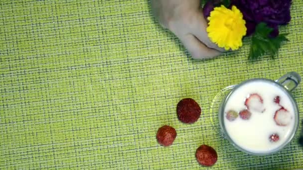Πάνω όψη, τραπέζι με μια κούπα γάλα και μούρα. υγιεινό πρωινό με φράουλες στο γάλα. Ένα μπουκέτο λουλούδια στο τραπέζι. Ένα χέρι βάζει ένα μπουκέτο από παιώνιες και μαργαρίτες σε ένα βάζο στο τραπέζι. — Αρχείο Βίντεο