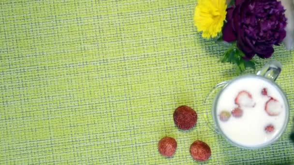 上のビュー,ミルクとベリーのマグカップとテーブル.ミルクの中のイチゴと健康的な朝食。テーブルの上に花の花束。手は牡丹とマリーゴールドの花の花瓶を移動します. — ストック動画