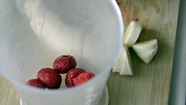 De hand doet stukjes fruit in de mengkom. Gesnipperde schijfjes aardbeien en banaan in een kom. Een vitaminesmoothie maken voor een dieetontbijt. Vruchtendrank. — Stockvideo