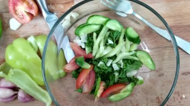 Bulgar biberi, salatalık, sarımsak ve otlu domatesler dilimlenip cam bir kasenin içine konuyor. Salata hazırlığı. Sağlıklı salata sebzeleri. Taze yıkanmış sebzeler masada.