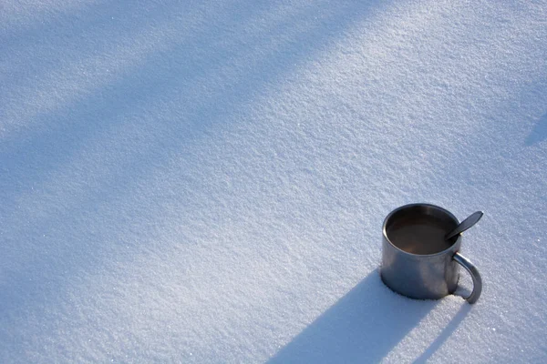 A mug in the snow. A metal cup with coffee stands in a snowdrift on a winter day. Shadows from trees on a snowy surface.