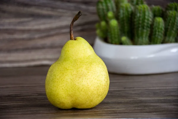 Pear on a wooden background. A yellow pear lies on the table against a blurred background. A cactus in a vase is blurred in the background.