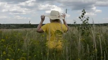 Çiçeklerin arasında şapkalı bir kadın. Yaşlı bir kadın çiçek açan bir tarlada yürüyor. Çayır bitkileri ve çiçekler.