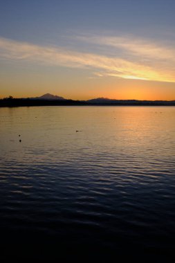 Baker Dağı ve Üç Kız Kardeş ufukta güneş ufuktan yükselirken görünmeye başlar. Öndeki mürekkepli maviden dağlara yakın turuncu ışığa sakin deniz geçişleri. Beyaz Kaya, BC, Kanada