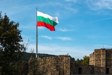 Bulgarian flag well illuminated with late afternoon sunshine flies from flagpole over ancient stone walls of Tsarevets Fortress, Veliko Tarnovo, Bulgaria