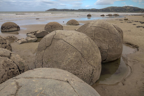 Large, barnacle encrusted, spherical boulders sit on sand of Koekohe Beach on the south-eastern coast of New Zealand's South Island.  Small tide pools have formed in the sand around them.