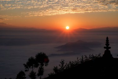 Güneş nihayet Himalaya tepelerinin üzerinde belirir ve puslu ön plana ışık ışınları yayar. Ön planda küçük bir şapşal silueti.