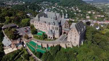 Yeşilliklerle çevrili tarihi bir kalenin havadan görünüşü ve arka planda bir şehir manzarası..