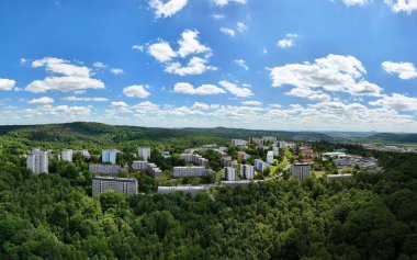 Mavi gökyüzünün altında yemyeşil ormanlarla çevrili bir şehrin havadan görünüşü.