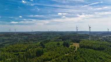 Mavi gökyüzünün altında yemyeşil bir arazi üzerindeki rüzgar türbinlerinin havadan görünüşü.