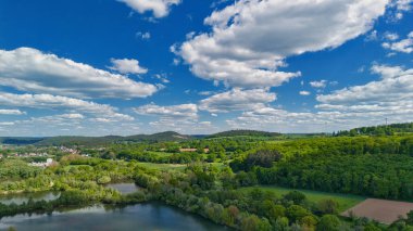 Ağaçları, tarlaları ve mavi gökyüzünün altında dağınık bulutları olan yemyeşil bir arazinin havadan görünüşü..