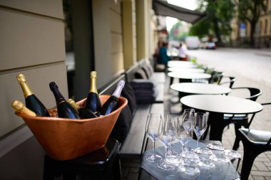 Şampanya şişeleri ve bakır buz kovasında köpüklü şarap ve yakında boş masa ve restoran terasında sandalyeler olan bardaklar.