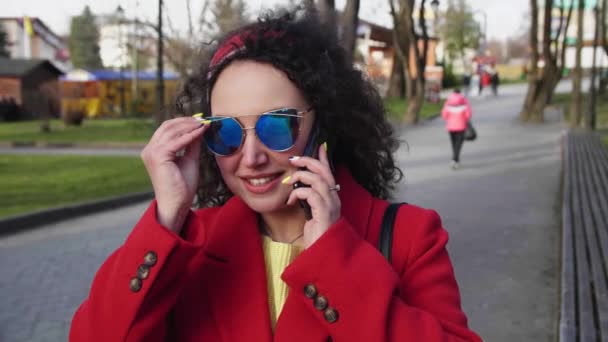 빨간 코트를 입고 선글라스를 낀 예쁘고 곱슬곱슬 한 아가씨 가 시내에 있는 스마트폰에서 — 비디오